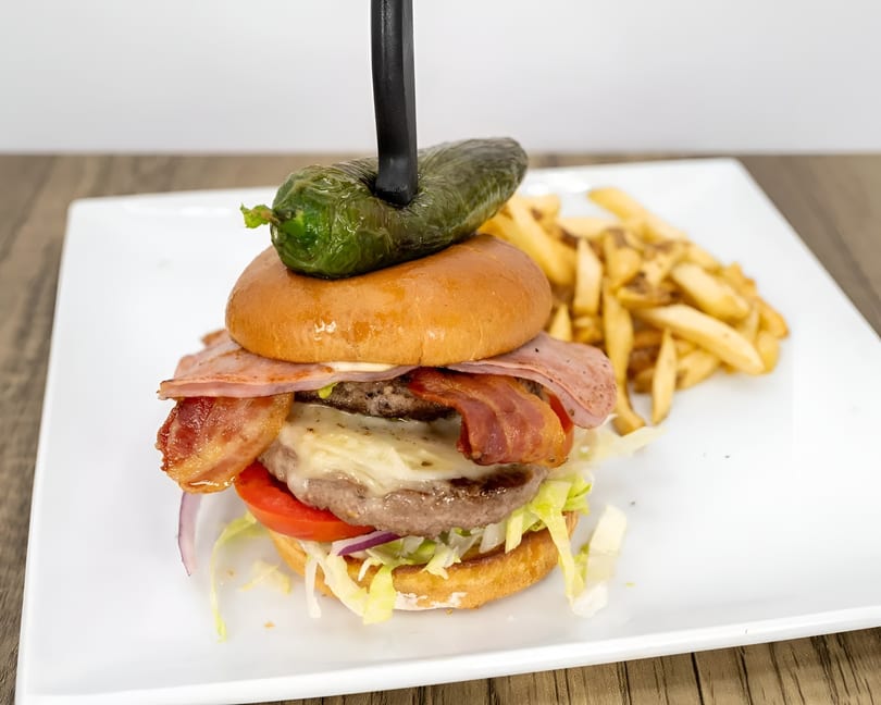 A large burger with bacon, ham, cheese, and vegetables, topped with a jalapeño and served with fries.