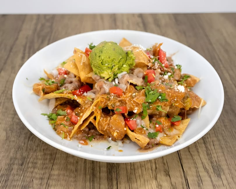 A white plate of nachos topped with guacamole, cheese, tomatoes, and cilantro on a wooden table.