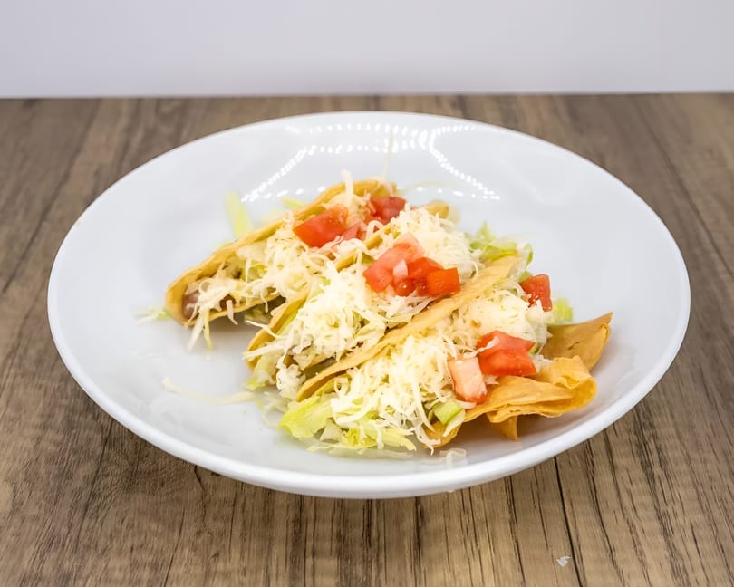 Three crispy tacos filled with meat, lettuce, shredded cheese, and diced tomatoes on a white plate.
