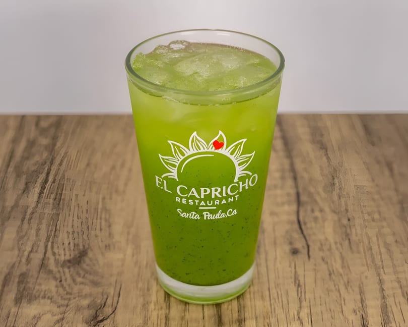 A tall glass of green iced beverage with the El Capricho Restaurant logo on a wooden table.