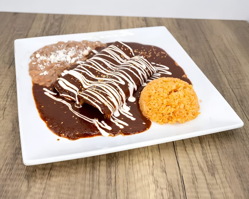 Three enchiladas covered in dark mole sauce with white crema, served with rice and refried beans.